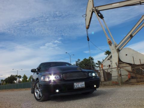 2003 Ford Crown Victoria - Dramatic low-angle photo of a black sedan with lit halo lamps and EVIL VIC plate against a cloudy sky and oil pump jack.