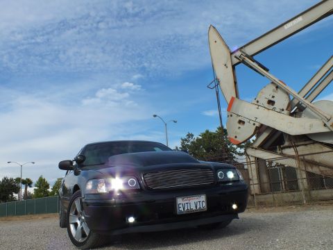 2003 Ford Crown Victoria - black sedan with glowing halo headlights and a California EVIL VIC plate sits on pavement beside a weathered oil pump jack.