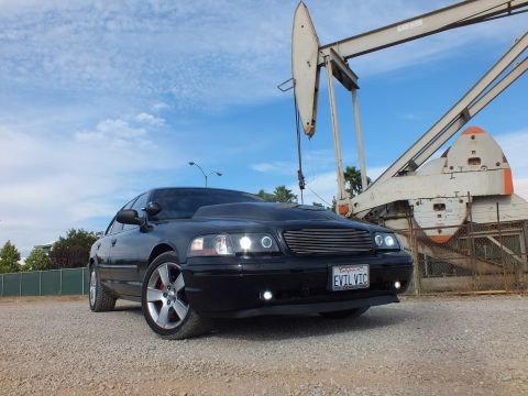 2003 Ford Crown Victoria - Front three-quarter view of a black sedan with halo headlights, billet grille and EVIL VIC plate in front of an oil pump jack.