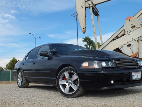 2003 Ford Crown Victoria - Front three-quarter shot of a glossy black sedan with chrome wheels and EVIL VIC plate in front of an oil pump jack.