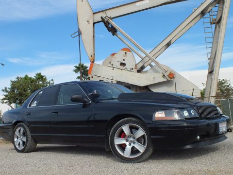 2003 Ford Crown Victoria - Side profile of a lowered black sedan with chrome five-spoke wheels parked beside a tall industrial oil pump jack.
