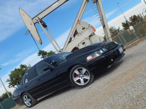 2003 Ford Crown Victoria - black sedan with billet grille shot at a dutch angle on gravel, a weathered oil pump jack filling the background.