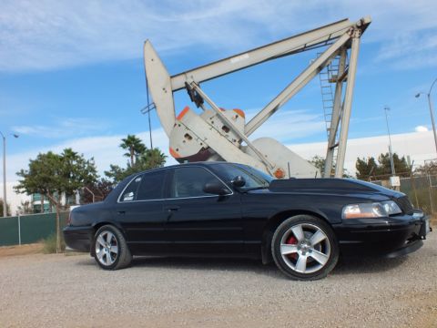 2003 Ford Crown Victoria - Side view of a lowered black sedan with chrome wheels on gravel, dwarfed by a large industrial oil pump jack.