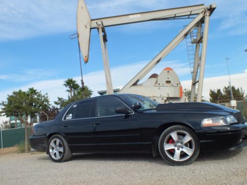 2003 Ford Crown Victoria - Side profile of a glossy black sedan with chrome five-spoke wheels parked before a weathered oil pump jack and palm trees.