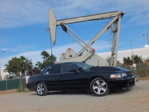 2003 Ford Crown Victoria - Side view of a lowered black sedan with chrome wheels on gravel, a tall oil pump jack and a bird in the sky behind.