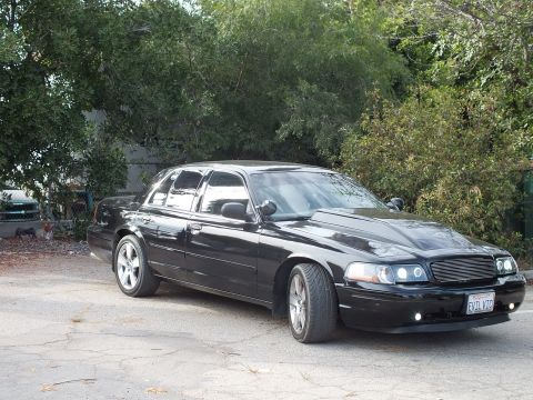 2003 Ford Crown Victoria - black sedan with billet grille, halo headlights and a California EVIL VIC plate parked on pavement beneath leafy trees.