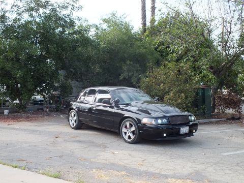2003 Ford Crown Victoria - glossy black sedan with chrome wheels and billet grille sits on cracked pavement surrounded by green foliage.