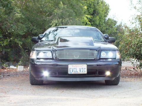 2003 Ford Crown Victoria - Head-on shot of a black Ford Crown Victoria with cowl hood and glowing halo headlights, EVIL VIC plate centered on its bumper.