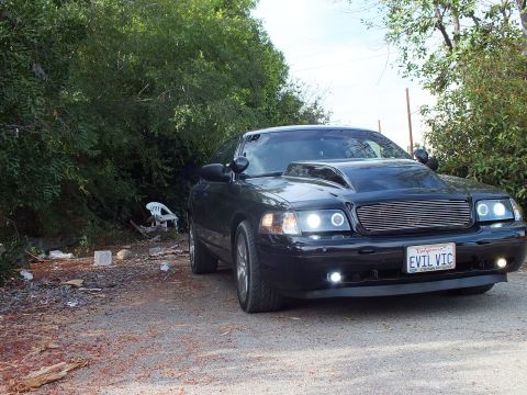 2003 Ford Crown Victoria - black Ford Crown Victoria with cowl hood, halo headlights and EVIL VIC plate parked on dirt beside roadside brush.
