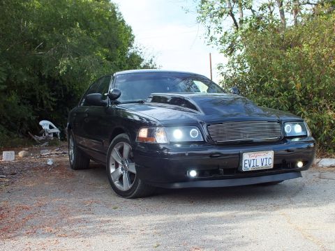 2003 Ford Crown Victoria - Front three-quarter view of a black Ford Crown Victoria with cowl hood, halo headlights and EVIL VIC plate amid greenery.