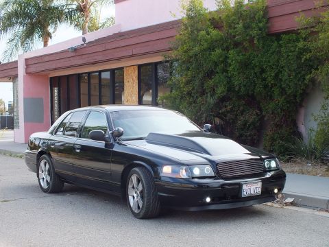 2003 Ford Crown Victoria - black full-size sedan with cowl hood and billet grille parked at a front-three-quarter angle outside a pink stucco building.