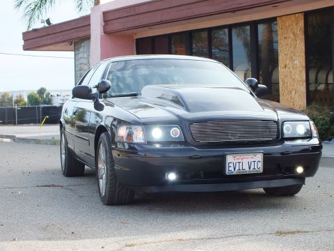 2003 Ford Crown Victoria - black full-size sedan with cowl hood and projector headlights lit, parked at a front-quarter angle beside a pink building and palm trees.