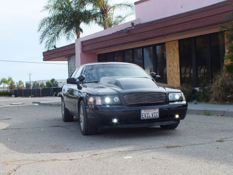 2003 Ford Crown Victoria - low front-three-quarter shot of a black full-size sedan with cowl hood, billet grille, and EVIL VIC plate, parked outside a pink building.
