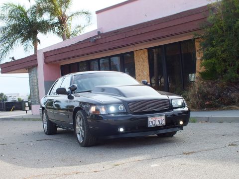 2003 Ford Crown Victoria - black full-size sedan with cowl hood and billet grille shown at a front-three-quarter angle on pavement beside a pink building and palm tre…