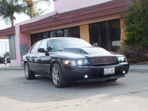 2003 Ford Crown Victoria - black full-size sedan with cowl hood, billet grille, and EVIL VIC plate parked at a front-quarter angle in front of a pink building.