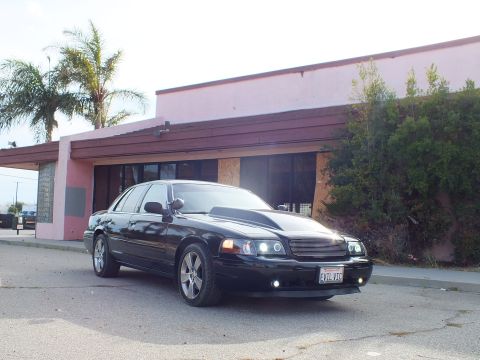 2003 Ford Crown Victoria - black full-size sedan with cowl hood and billet grille parked at a front-quarter angle in warm evening light beside a pink building.