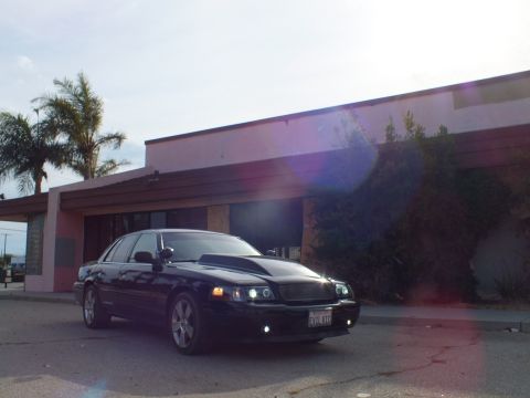 2003 Ford Crown Victoria - black full-size sedan with cowl hood parked at a front-quarter angle outside a pink building, with sun lens flare across the frame.