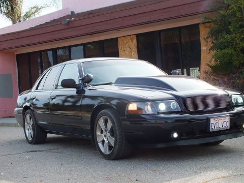 2003 Ford Crown Victoria - black full-size sedan with cowl hood, billet grille, and lit projector headlights parked at a front-quarter angle outside a pink building.