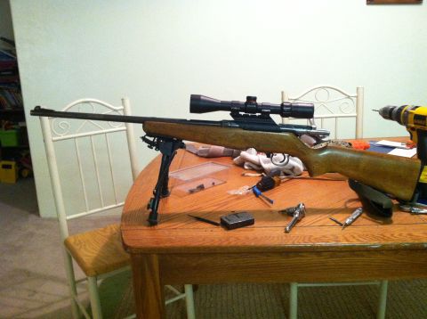1989 Ford Bronco - A rifle with a scope and bipod rests on a table next to tools and a magazine.