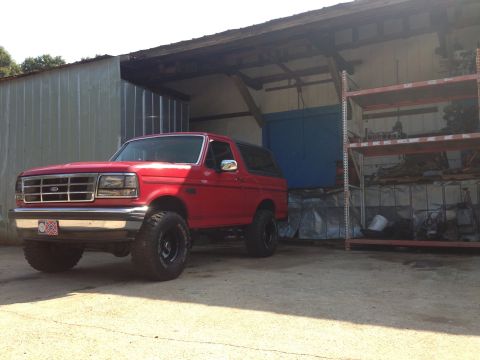 1992 Ford Bronco - A red 1992 Ford Bronco with aftermarket wheels is parked in front of a garage with shelves of parts.
