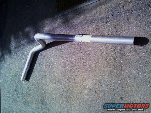 1982 Ford Bronco - A metal exhaust pipe with a distinct bend lies on a gravel surface, showing a label and a black tip.
