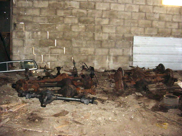 Several rusty vehicle axles and parts scattered on a dirt floor in front of a cinder block wall