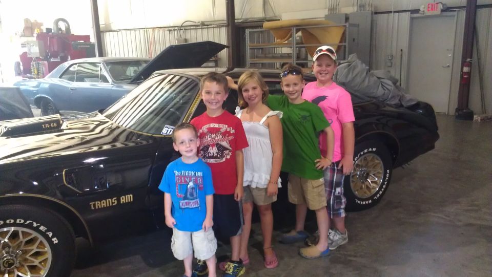 Kids pose in front of a black Pontiac Trans Am. Another car is in the background