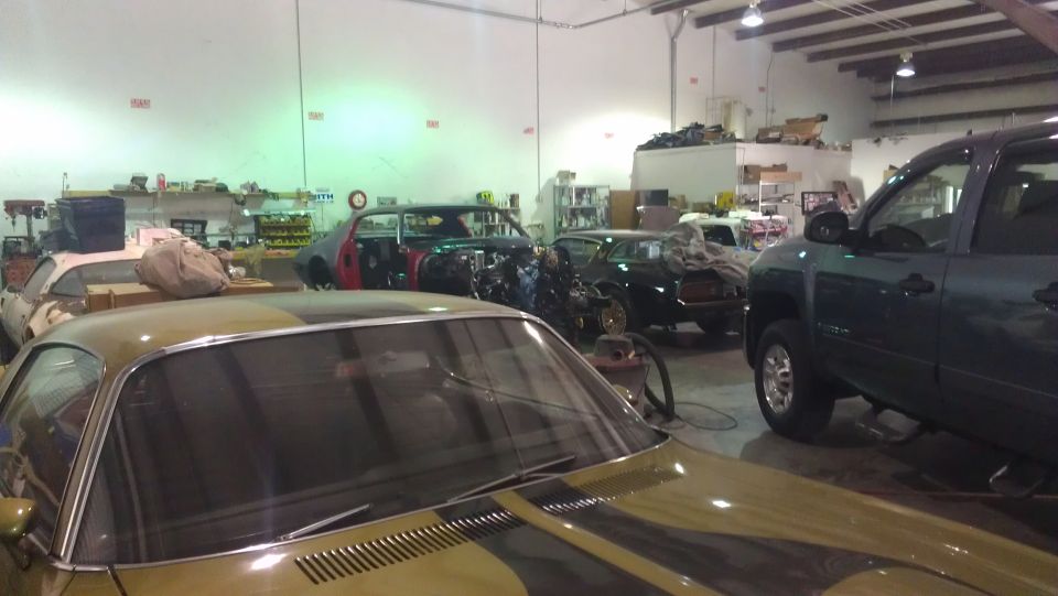 Gold car with black stripes in foreground, garage with several cars and a truck