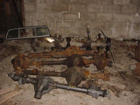 1992 Ford Bronco - Multiple vehicle axles are stored in a cluttered, dimly lit room with a concrete block wall.