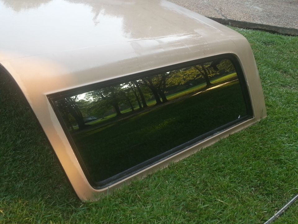 Tan truck cap with a tinted window sitting on grass. Trees and sky reflected in the window