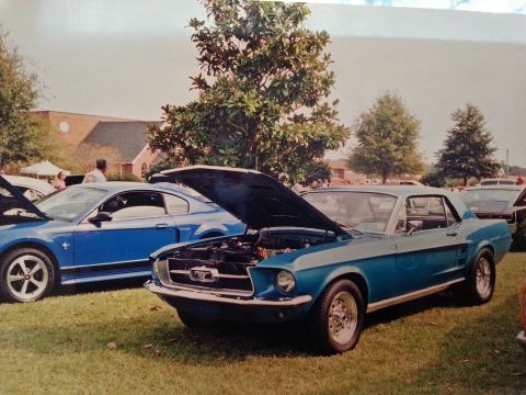 1967 Ford Mustang - A classic 1967 Ford Mustang is displayed with its hood up at a car show, alongside a more modern Mustang model.