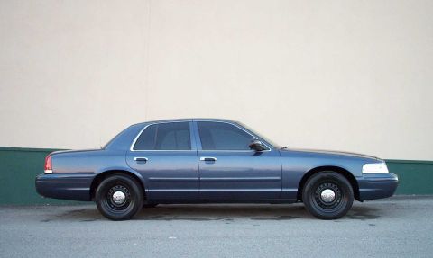 1998 Ford Crown Victoria - Side view of a blue sedan with black steel wheels and tinted windows parked against a light-colored wall.