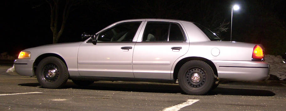 Side view of a silver Crown Victoria sedan with black steel wheels parked at night under a streetlight