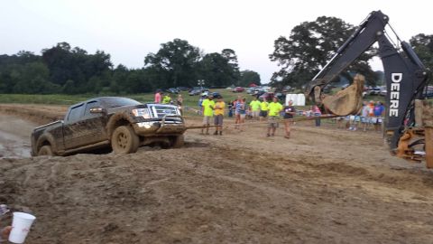 1987 Ford Bronco - Muddy Ford F-150 being pulled from a pit by a Deere excavator, with onlookers present.