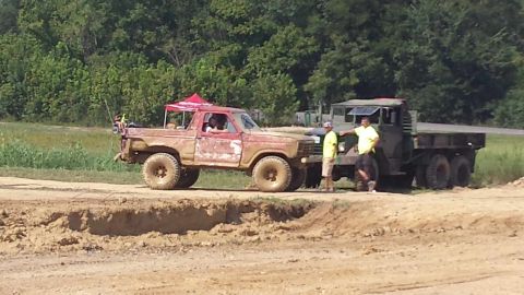 1987 Ford Bronco - A red pickup truck sits off-road next to a large military truck on a sunny day.