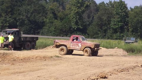 1987 Ford Bronco - Muddy red pickup truck is being towed by a military flatbed truck on a dirt track.