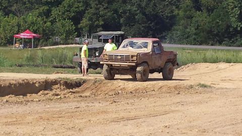 1987 Ford Bronco - Two trucks sit in a dirt lot, one appears to be stuck or recovering from off-roading.