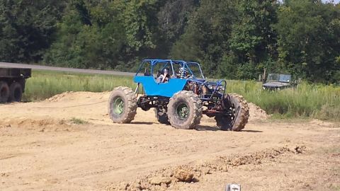 1987 Ford Bronco - A custom-built blue buggy with oversized tires navigates a dirt track, with a trailer in the background.