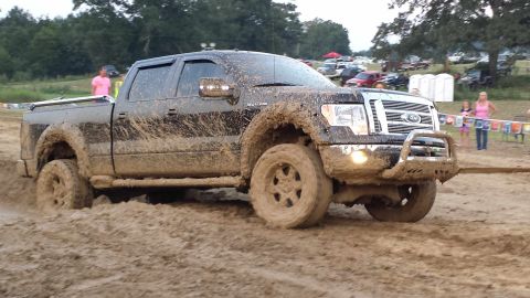 1987 Ford Bronco - A black pickup truck is covered in mud as it navigates a muddy track, with spectators and other vehicles visible in the distance.