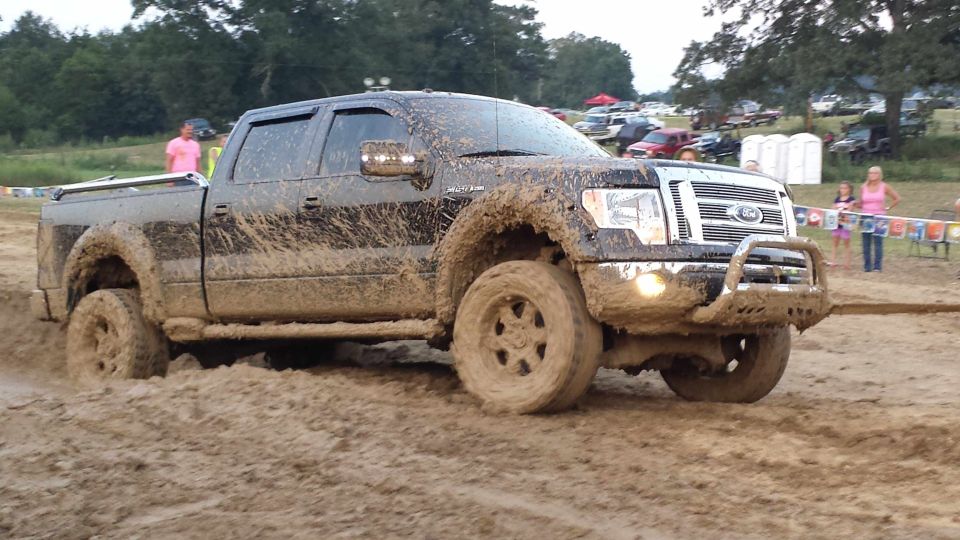 Muddy pickup truck driving through a dirt track with people and other vehicles in the background