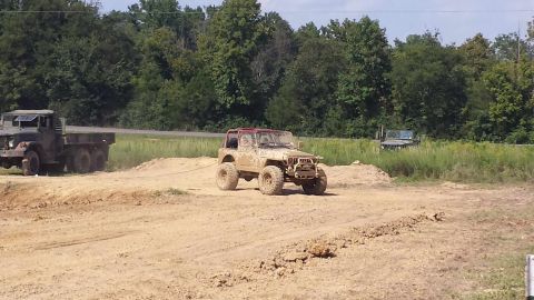 1987 Ford Bronco - A muddy off-road vehicle is being towed by a large military truck in a dirt area.
