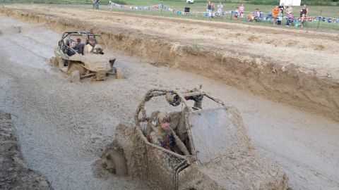 1987 Ford Bronco - Two off-road vehicles navigate a muddy track during a competition, with spectators watching from the sidelines.