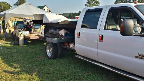 1987 Ford Bronco - White pickup truck with a flatbed and dual rear wheels is parked on grass, towing a trailer.