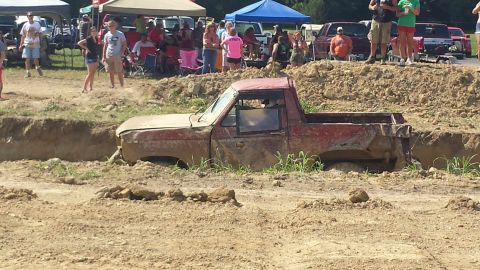 1987 Ford Bronco - Pickup truck stuck in mud pit during an event, with onlookers in the background.