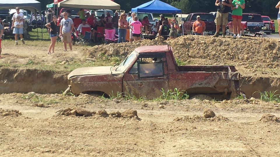 Red pickup truck stuck in a mud pit, with spectators watching from higher ground