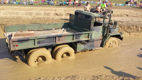 1987 Ford Bronco - A military flatbed truck is stuck deep in a mud pit during an off-road event, with spectators in the background.