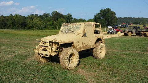 1987 Ford Bronco - A heavily mud-caked Jeep Wrangler sits in a grassy field with other vehicles in the background.