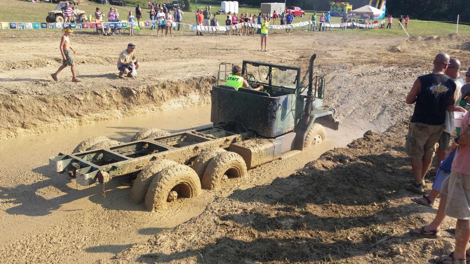 Military truck chassis with multiple axles stuck deep in mud. Driver wearing a green 'STAFF' shirt is visible in the cab. Spectators watch from the bank
