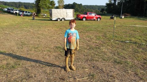 1987 Ford Bronco - Boy covered in mud stands in a field with vehicles and a trailer in the background.