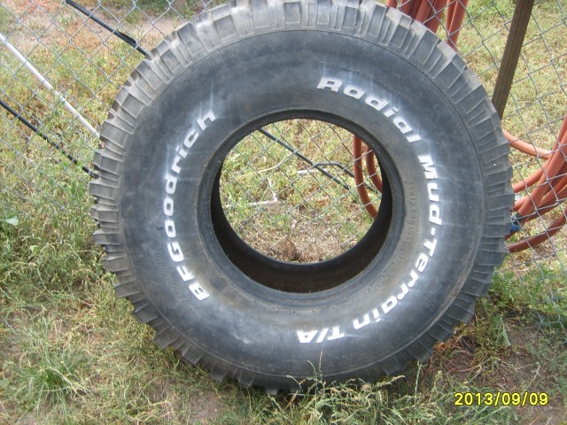 BFGoodrich Mud-Terrain T/A tire with white lettering, mounted on a rim, resting on grass near a chain-link fence